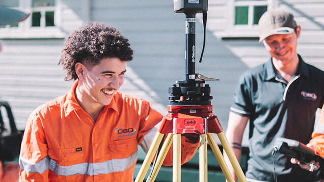 Two people stand outdoors using surveying equipment; one wears an orange safety uniform and smiles, while the other wears a dark polo shirt and holds a device.