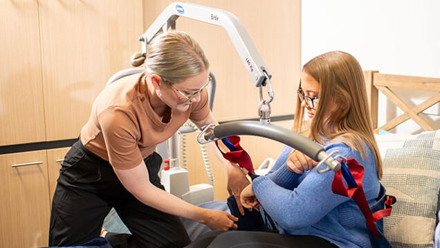 A caregiver assists a seated woman with a ceiling hoist in a home or clinical setting.