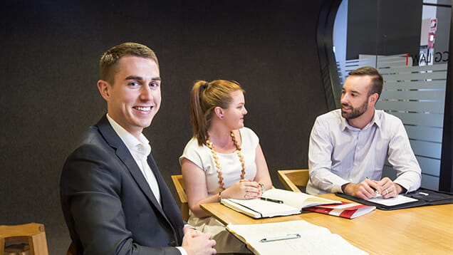 Three people in business attire sit at a table with notebooks and folders, having a discussion in a modern office setting. One man smiles at the camera.