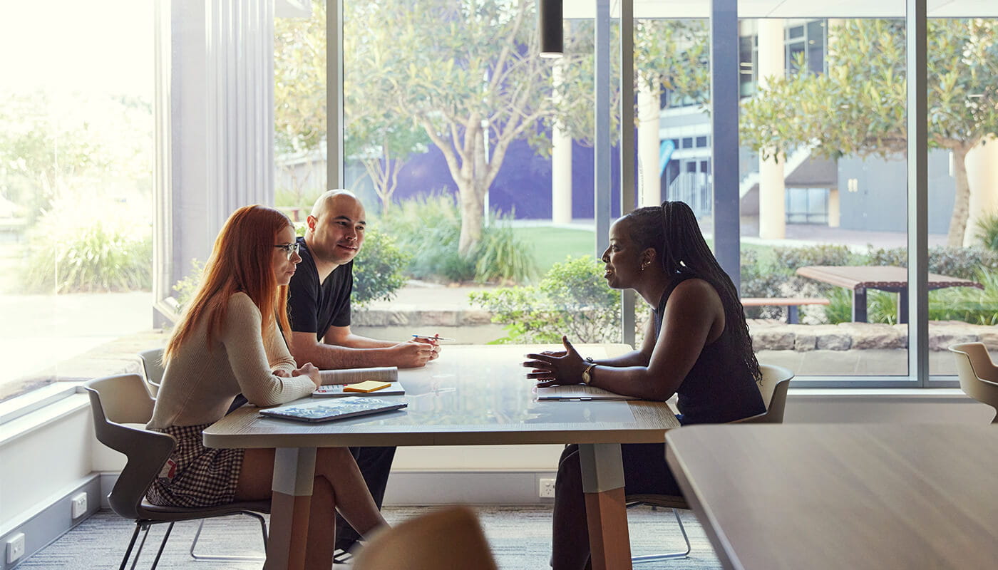 Three people sit at a table in a modern office, engaged in conversation with notebooks and pens, next to large windows overlooking trees and benches outside.