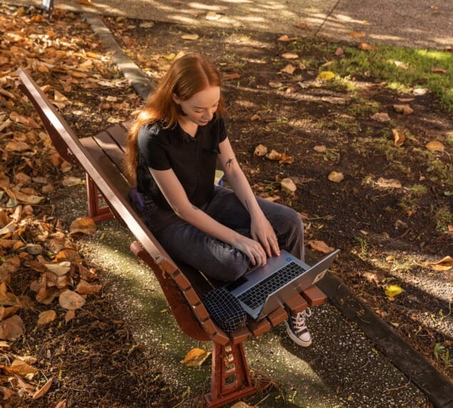 A person with long red hair sits on a park bench, working on a laptop, surrounded by fallen leaves and partial sunlight.