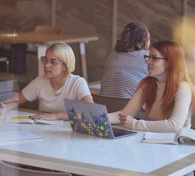 Two women sit at a table with notebooks and a laptop covered in a floral design, engaged in discussion in a classroom or study setting.