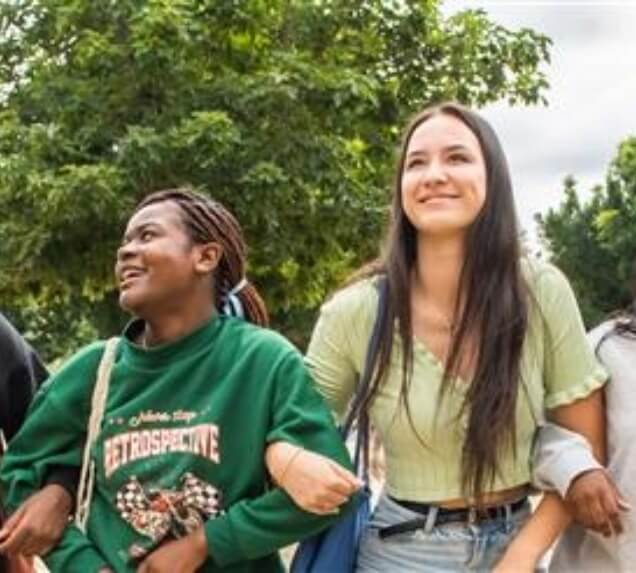 Two young women walking outdoors, smiling and linking arms, with trees in the background on a cloudy day.
