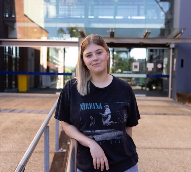 A young woman with straight, blonde hair stands outdoors in front of a modern glass building, wearing a black Nirvana t-shirt and looking at the camera.