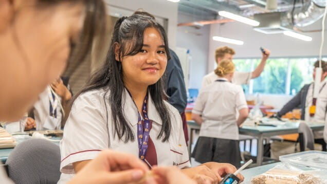 A student in a white uniform sits at a classroom table, smiling at the camera while others work in the background.