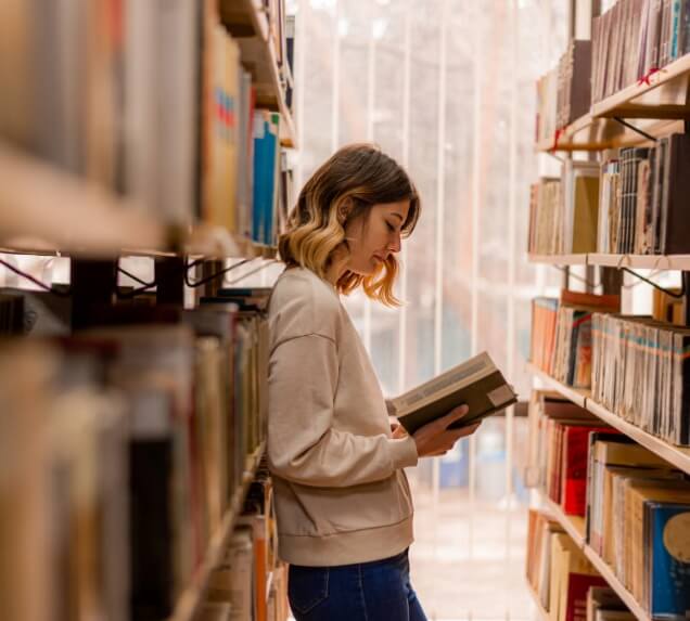 A woman stands between bookshelves in a library, reading a book while leaning against a shelf.