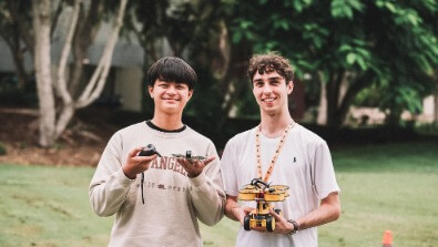 Two young men standing outdoors, holding a small drone and a remote control, with trees and grass in the background.