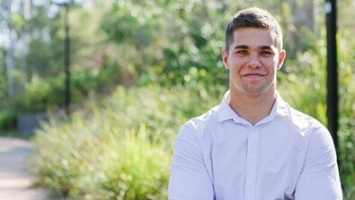 A young man with short hair wearing a white shirt stands outdoors with greenery in the background, smiling slightly.