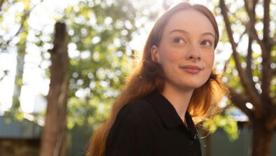 A young person with long reddish hair and a nose ring stands outdoors, smiling slightly, with sunlight and trees in the background.