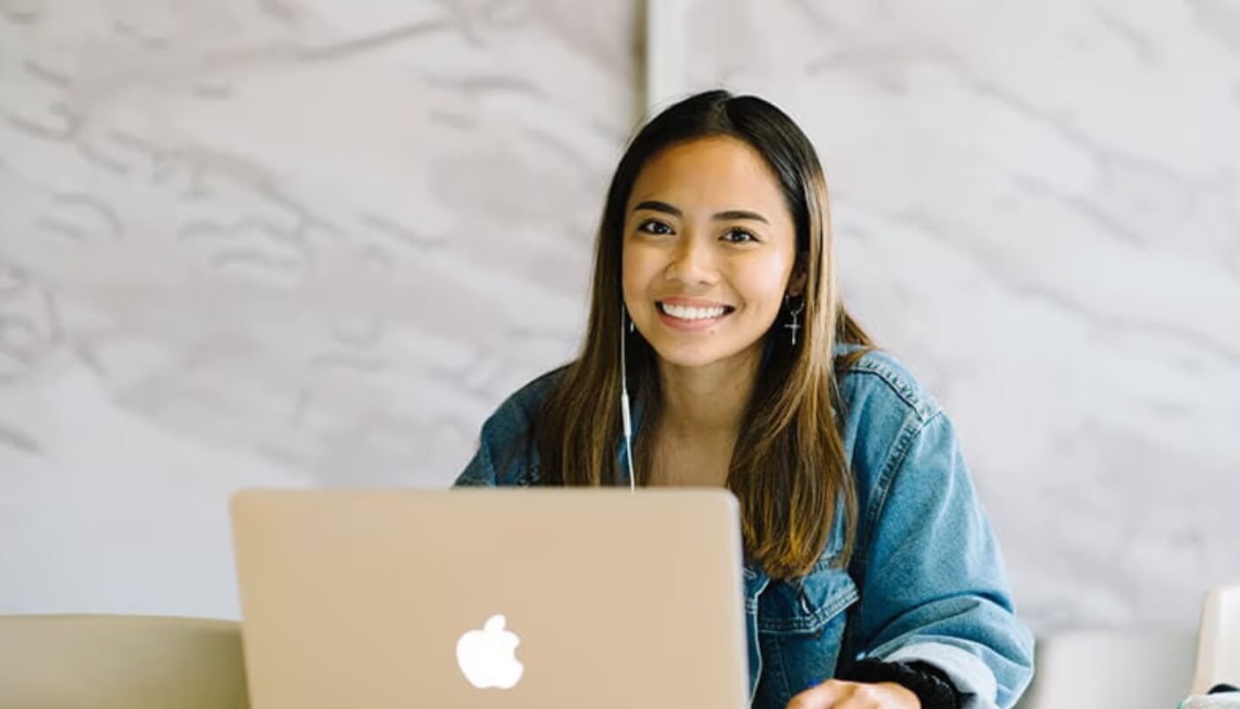 A person with long dark hair, wearing a denim jacket, sits at a table smiling while using a silver Apple laptop.