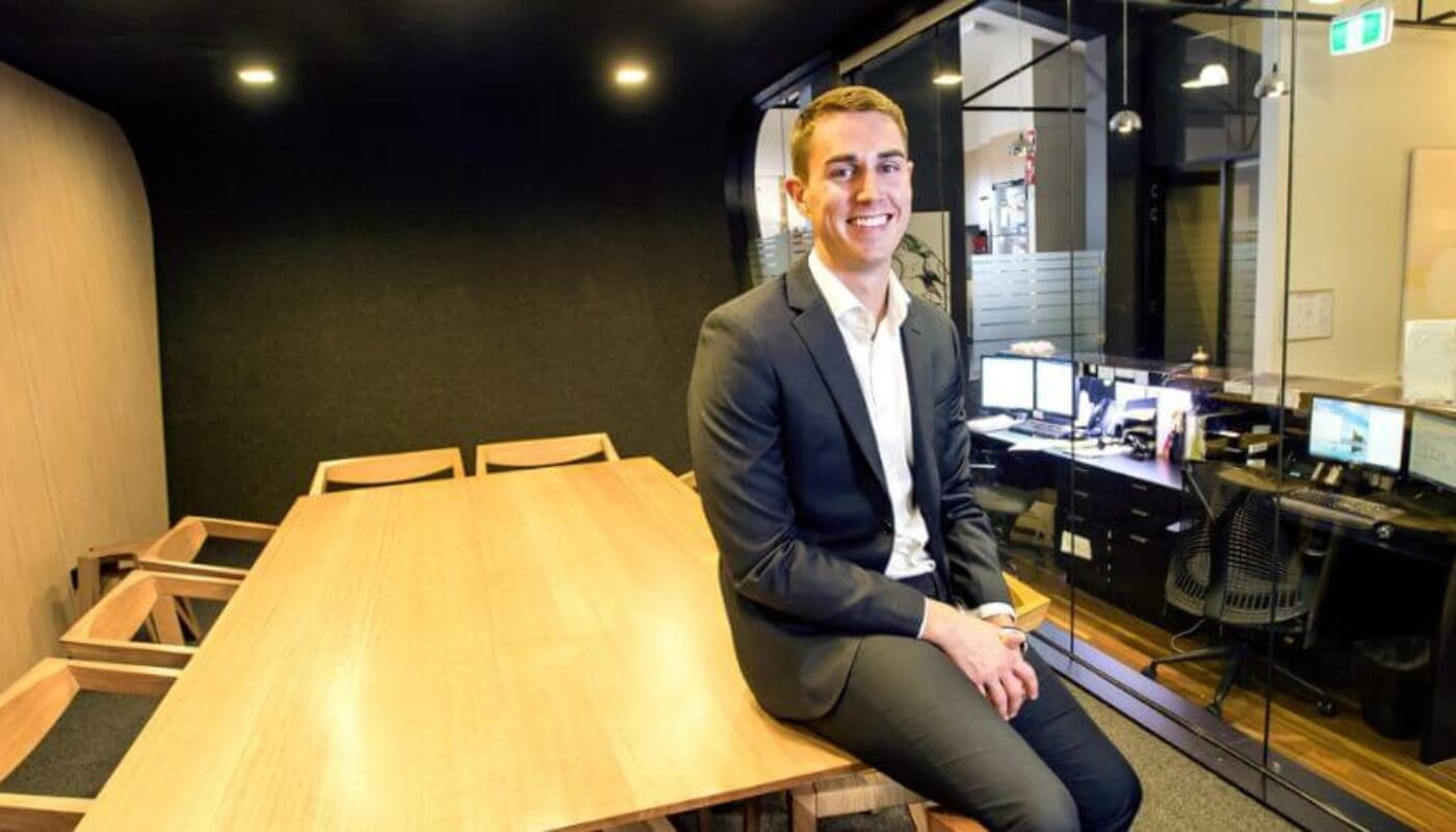 A man in a suit sits on the edge of a wooden conference table in a modern office with glass walls and computer workstations in the background.