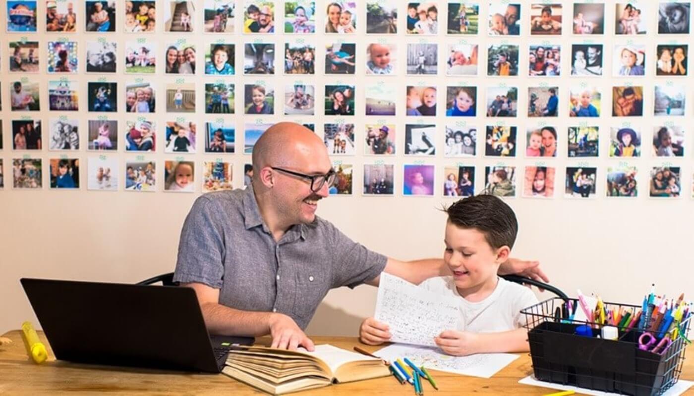 An adult and child sit at a table with books and papers, smiling while working together. A wall behind them displays a grid of family photographs.