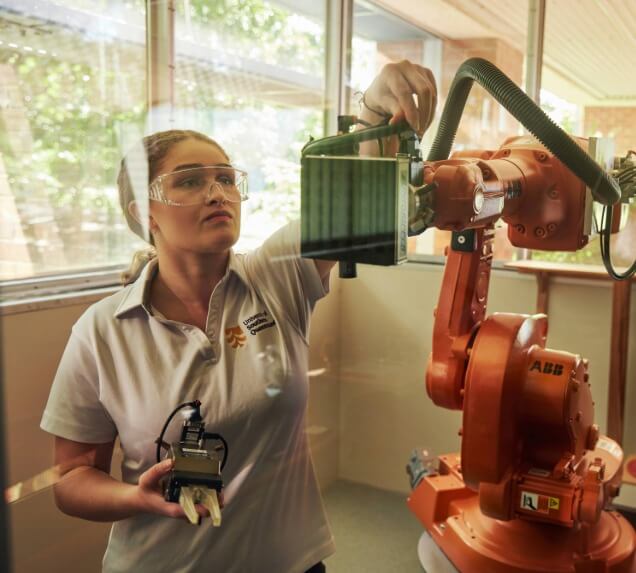 A woman wearing safety glasses operates an orange industrial robotic arm in a lab, holding a tool and adjusting a component.