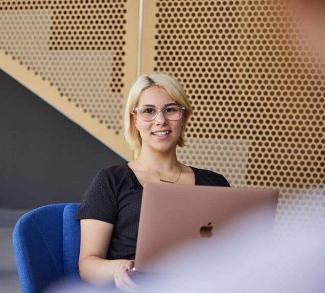 A person with short blonde hair and glasses sits on a blue chair, smiling while using a laptop in front of a modern perforated wall.