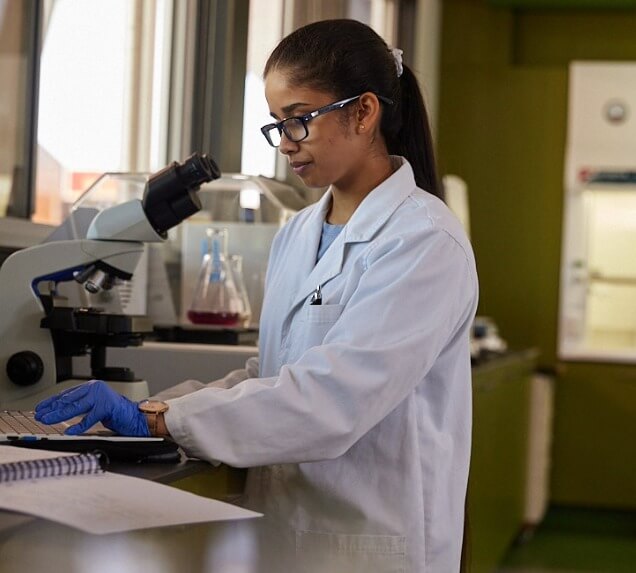 A woman in a lab coat and gloves uses a computer in a laboratory, with a microscope and lab equipment visible on the table beside her.