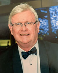An older man with light hair and glasses is wearing a tuxedo and bow tie, standing indoors with blue and yellow lights in the background.