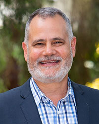 A middle-aged man with short gray hair and a beard, wearing a blue blazer over a checkered shirt, smiling outdoors.