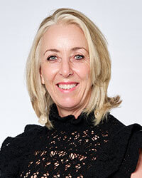 A woman with shoulder-length blonde hair wearing a black lace top smiles at the camera against a plain background.