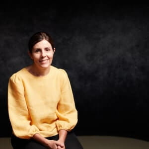 Woman with dark hair in a low bun, wearing a yellow long-sleeve blouse, sits and smiles at the camera against a dark, plain background.