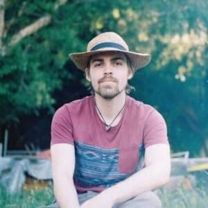 A man with a beard and mustache, wearing a straw hat and a red t-shirt, sits outdoors on grass with trees and sunlight in the background.