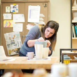 A woman in a striped shirt works on shaping a ceramic piece in a pottery studio, with several finished pieces and art supplies on the table.