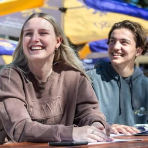 Two young people sit at a table outdoors, smiling and looking in the same direction. Sunny weather with yellow and blue umbrellas in the background.