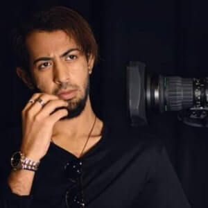 A man with a beard and mustache poses thoughtfully in front of a professional camera against a dark background.