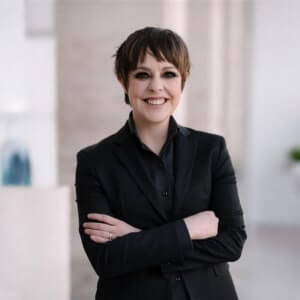 A person with short brown hair, wearing a black suit, stands indoors with arms crossed and smiles at the camera.