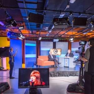 A TV studio with cameras, lights, two people seated for an interview, crew members working, and a monitor displaying a singer's close-up.