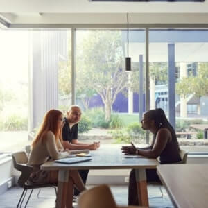 Three people sit at a table in a modern office space, engaged in discussion near large windows with trees visible outside.