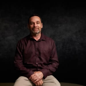 A man with short hair and a beard sits on a stool against a dark, textured background, wearing a maroon shirt and light-colored pants.