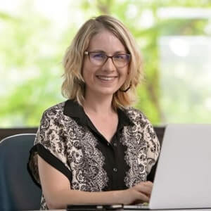 A woman with blonde hair and glasses sits at a desk, smiling at the camera while working on a laptop. A smartphone is on the desk. The background is bright and blurred with greenery.