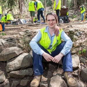 A person wearing a yellow safety vest and work boots sits on rocks in a wooded area, with several people in safety vests working in the background.