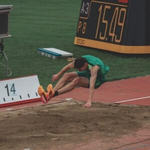 An athlete in green lands in a sandpit during a long jump event, with a scoreboard displaying 15.49 meters in the background.