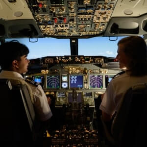 Two pilots sit in the cockpit of a commercial airplane, facing the controls and flight instruments, with a view of blue sky through the windows.