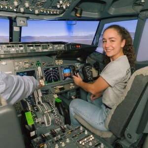 Young woman sitting in the pilot seat of an airplane cockpit, smiling at the camera, with another person’s arm visible adjusting controls.