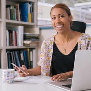A woman sits at a desk with papers, a pen, a laptop, and a mug, smiling at the camera. Bookshelves are visible in the background.