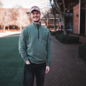 A man wearing a green sweater, dark jeans, and a cap stands on a brick pathway next to a grassy area, with buildings and trees in the background.