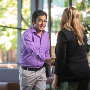 A man in a purple shirt and a woman in a black jacket are shaking hands indoors, with blurred greenery visible through the windows behind them.