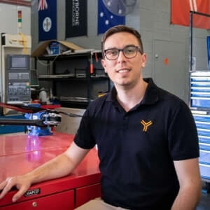 A man with glasses sits at a red workbench in a workshop, with tools, equipment, and flags visible in the background.