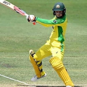 A cricket player in a yellow and green uniform, wearing a helmet, prepares to hit the ball with a bat during a match on a grassy field.