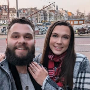 A man with a beard and a woman in a plaid coat stand close together and smile at the camera, with a canal and buildings in the background.
