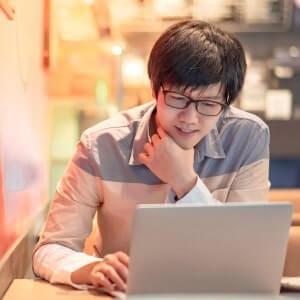 Person with glasses sitting at a table and looking at a laptop screen, appearing focused in a brightly lit indoor setting.