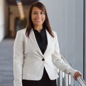 A woman in a white blazer and black shirt stands indoors by a glass railing, smiling at the camera in a modern hallway.