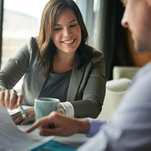 Two people sit at a table discussing documents; the woman smiles and holds a pen while the man points to a page.