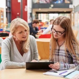 An adult woman and a teenage girl sit at a table in a library, looking at a tablet together. The girl has a notebook and pen on the table.