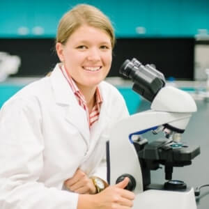 A person in a white lab coat sits next to a microscope in a laboratory, smiling at the camera.