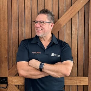A man wearing glasses and a black polo shirt stands in front of a wooden door with his arms crossed, smiling and looking to the side.