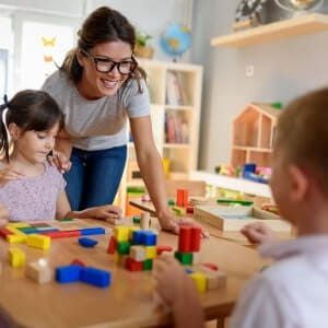 A teacher smiles while helping young children play with colorful wooden blocks at a classroom table.