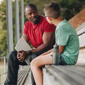 An adult in a red shirt holding a clipboard talks to a boy in a green shirt sitting on stadium bleachers.